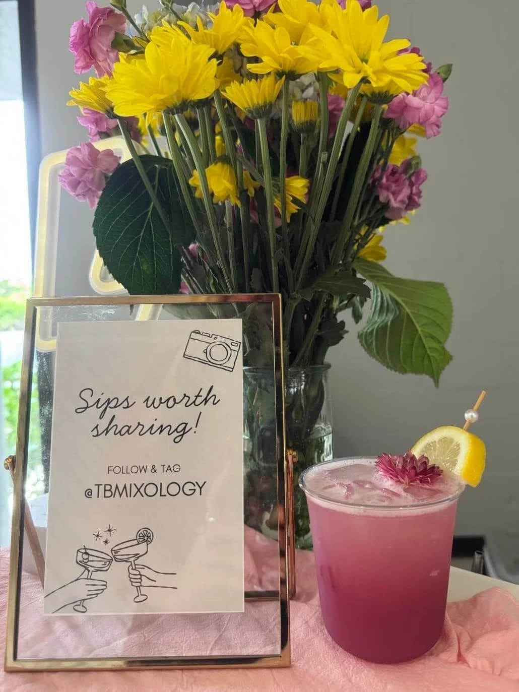 Sips Worth Sharing signage with fresh flowers and a violet cocktail