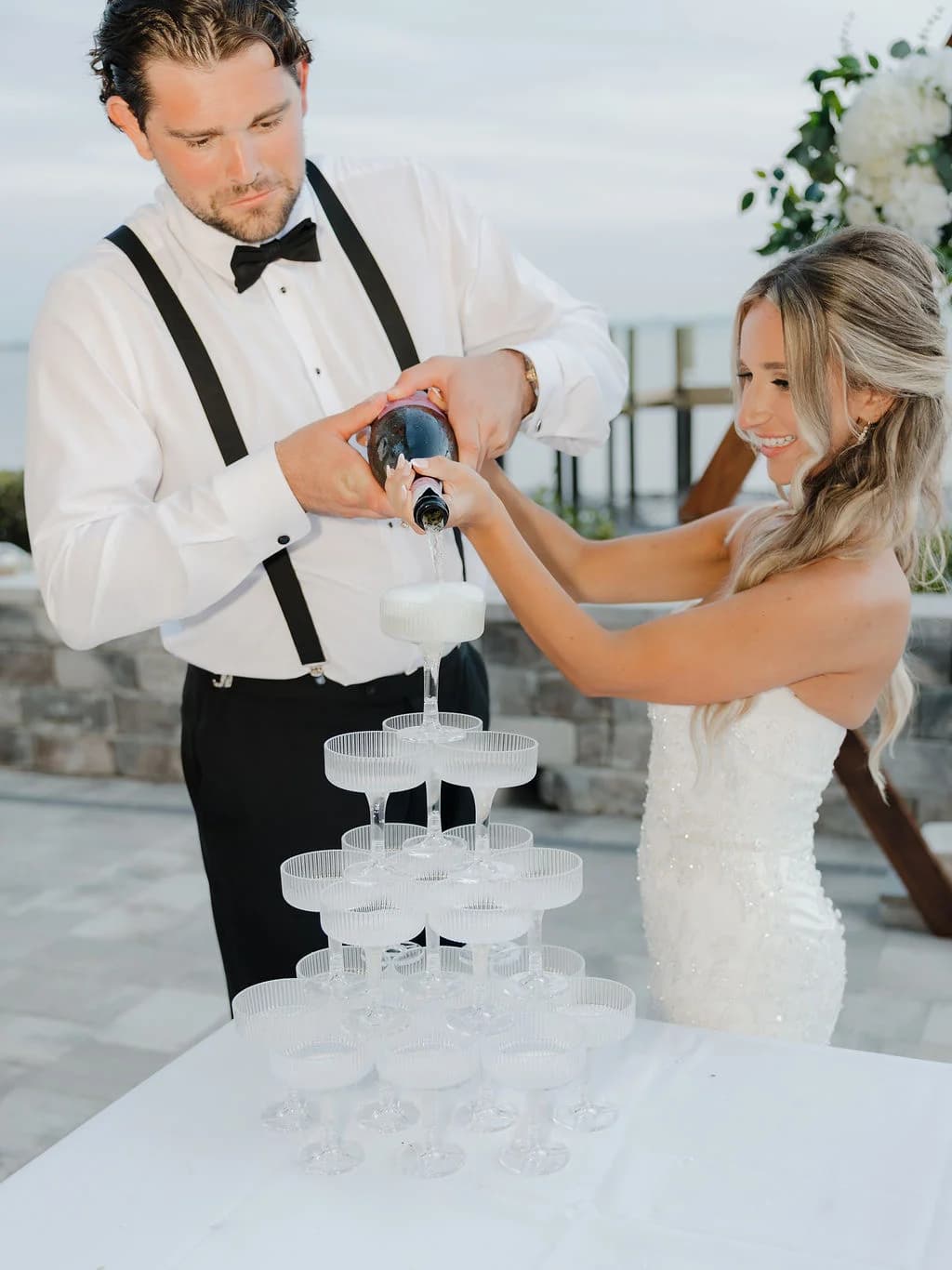 Wedding couple pouring a champagne tower beside a white floral arch